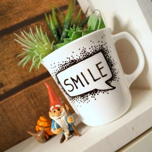 May include: White ceramic mug with black polka dots and the word "SMILE" written in a speech bubble. The mug is filled with small green plants and is sitting on a white shelf.