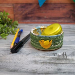 May include: A small, round, green and tan fabric-covered container with a yellow felt bird applique. Inside is a yellow object. A pencil, pen, and marker are to the left. Paper clips are to the right.