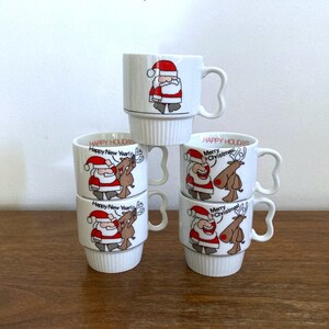 May include: Set of six white ceramic mugs with Christmas-themed illustrations. Each mug features Santa Claus and a reindeer with festive greetings like "Happy Holidays" and "Merry Christmas".