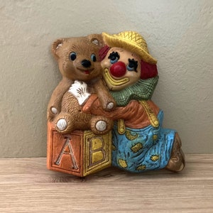May include: A ceramic figurine of a clown holding a teddy bear. The clown is wearing a yellow hat, a blue and green outfit, and has a red nose. The teddy bear is brown and has a white belly. The figurine is sitting on top of a block with the letters A and B on it.