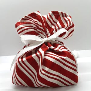 May include: A fabric pouch featuring a red and white diagonal striped pattern. The top of the pouch is gathered and secured with a white ribbon tied in a bow, creating a ruffled effect at the opening. The material appears to be a soft cotton textile.