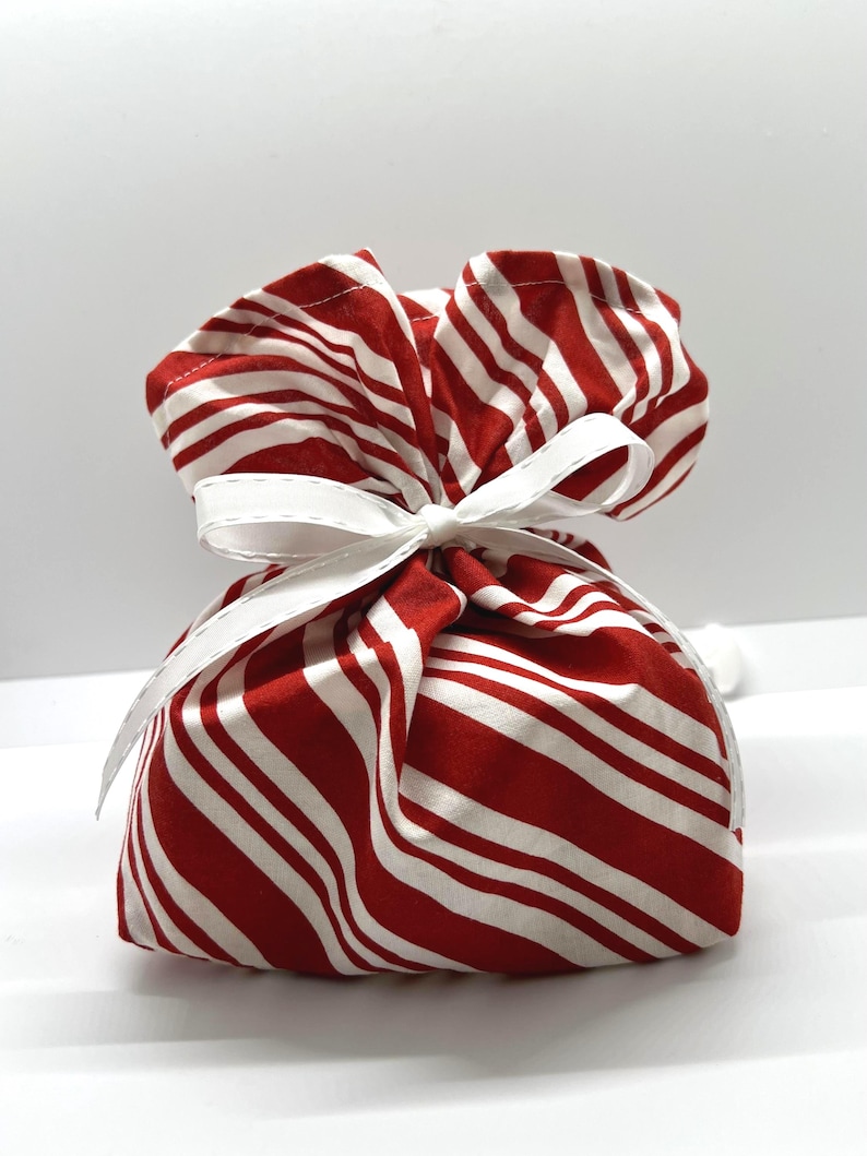 May include: A fabric pouch featuring a red and white diagonal striped pattern. The top of the pouch is gathered and secured with a white ribbon tied in a bow, creating a ruffled effect at the opening. The material appears to be a soft cotton textile.