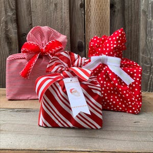 May include: Three fabric gift bags in red and white patterns. One bag features thin horizontal stripes with a red ribbon, one has red and white diagonal candy cane stripes with a tag showing a Santa illustration, and one has white polka dots.