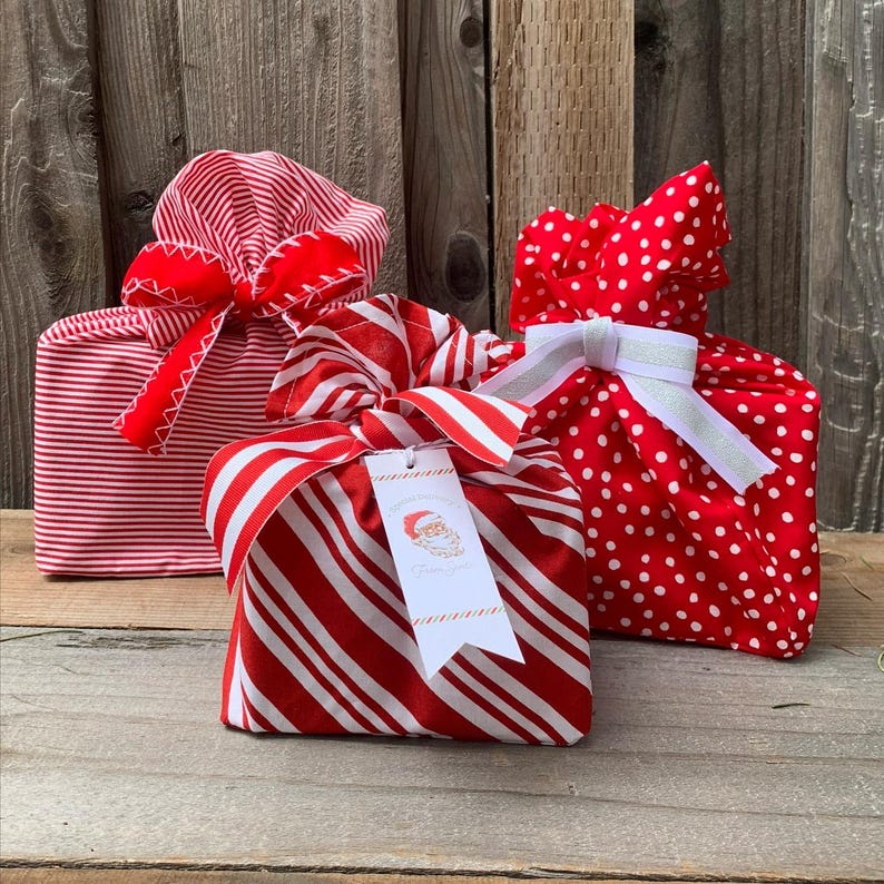 May include: Three fabric gift bags in red and white patterns. One bag features thin horizontal stripes with a red ribbon, one has red and white diagonal candy cane stripes with a tag showing a Santa illustration, and one has white polka dots.