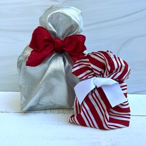 May include: A metallic silver fabric gift bag tied with a large red ribbon bow, positioned next to a smaller fabric gift bag featuring a red and white striped pattern and secured with a crisp white ribbon bow, both standing upright.