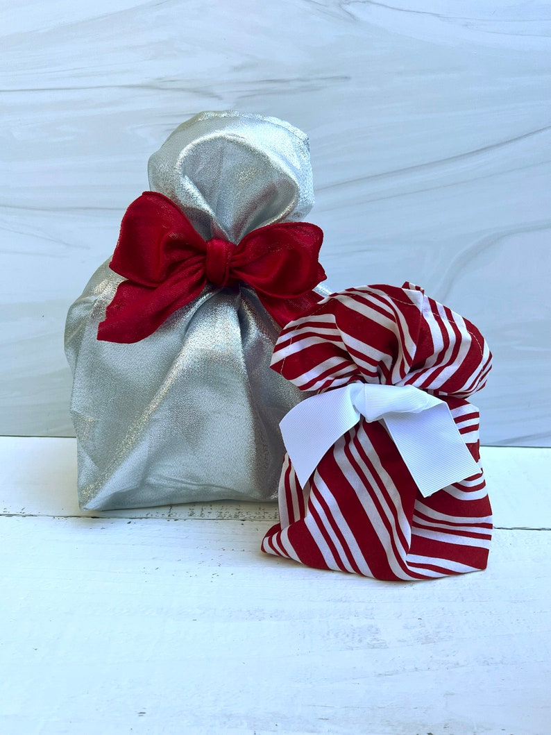 May include: A metallic silver fabric gift bag tied with a large red ribbon bow, positioned next to a smaller fabric gift bag featuring a red and white striped pattern and secured with a crisp white ribbon bow, both standing upright.