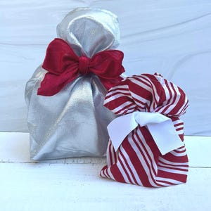 May include: Two fabric gift bags. The larger bag is made of a metallic silver fabric and is tied with a wide, deep red velvet ribbon. The smaller bag features a red and white candy cane striped pattern and is tied with a crisp white ribbon.