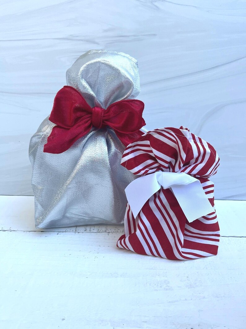 May include: Two fabric gift bags. The larger bag is made of a metallic silver fabric and is tied with a wide, deep red velvet ribbon. The smaller bag features a red and white candy cane striped pattern and is tied with a crisp white ribbon.
