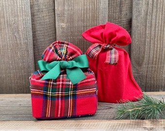 May include: Two fabric gift bags, one square-shaped with a red, green, and yellow tartan pattern tied with a solid green ribbon, and one taller, solid red drawstring bag tied with a matching tartan fabric ribbon, presented as a pair.