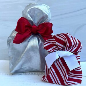 May include: A metallic silver fabric gift bag cinched at the top with a large red ribbon bow, positioned next to a smaller fabric gift bag featuring a red and white vertical striped pattern and a white ribbon bow tied around the gathered top.