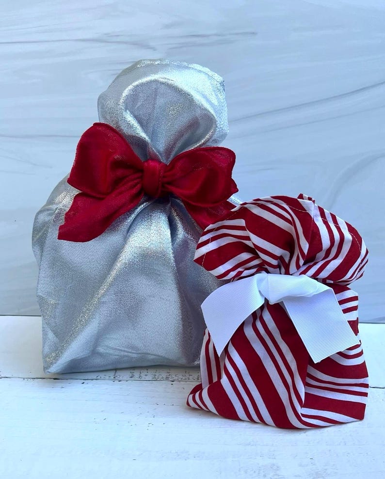 May include: A metallic silver fabric gift bag cinched at the top with a large red ribbon bow, positioned next to a smaller fabric gift bag featuring a red and white vertical striped pattern and a white ribbon bow tied around the gathered top.