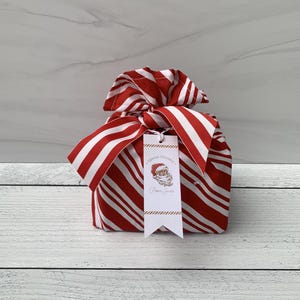 May include: A fabric gift bag featuring a red and white diagonal candy cane stripe pattern, tied at the top with a matching fabric bow. A rectangular white gift tag hangs from the bow, displaying a Santa Claus illustration and the text Special Delivery From Santa.