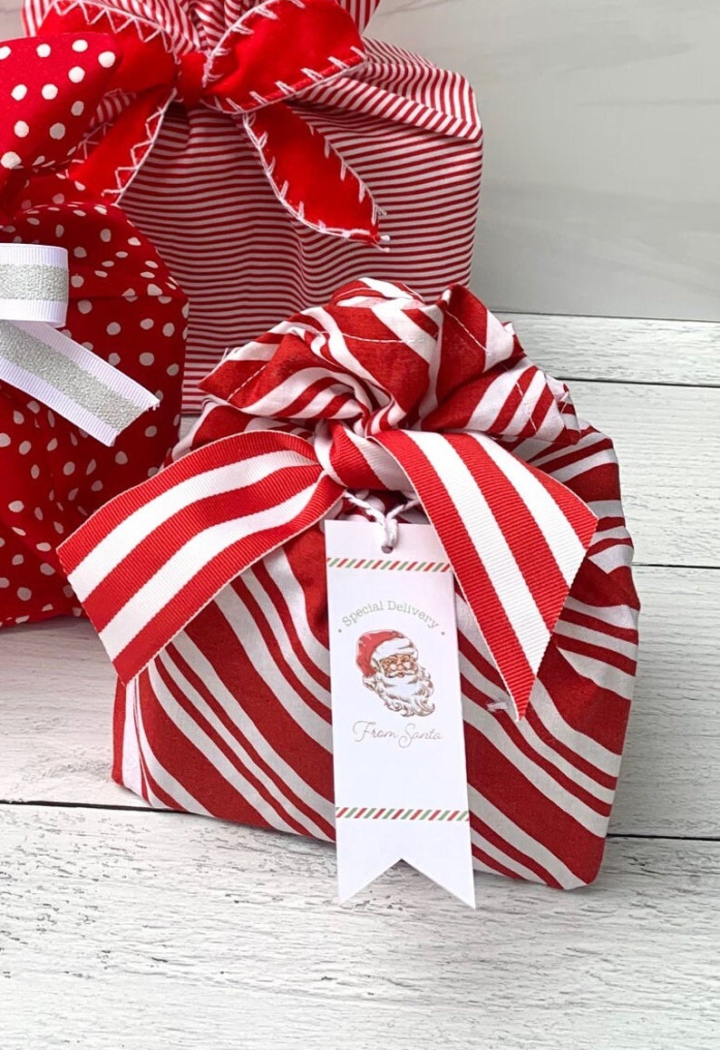 May include: A fabric gift wrap bundle featuring a red and white diagonal stripe pattern, tied with a matching striped ribbon. A white rectangular gift tag hangs from the ribbon, displaying a Santa Claus illustration and the text Special Delivery From Santa.