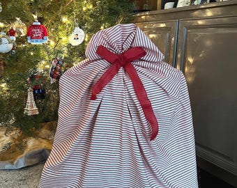 May include: A large fabric gift sack featuring a pattern of thin horizontal red and white stripes. The sack is gathered at the top and secured with a wide, solid red ribbon tied into a bow, creating a draped, voluminous shape for holding items.