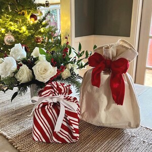 May include: A square gift bag featuring a red and white candy cane striped pattern tied with a white ribbon, positioned next to a plain cream-colored fabric gift bag cinched at the top with a large, vibrant red fabric bow.