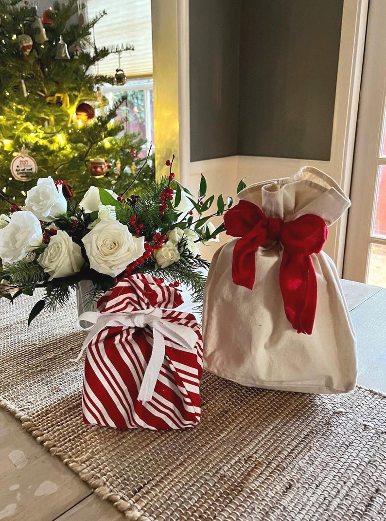 May include: A square gift bag featuring a red and white candy cane striped pattern tied with a white ribbon, positioned next to a plain cream-colored fabric gift bag cinched at the top with a large, vibrant red fabric bow.