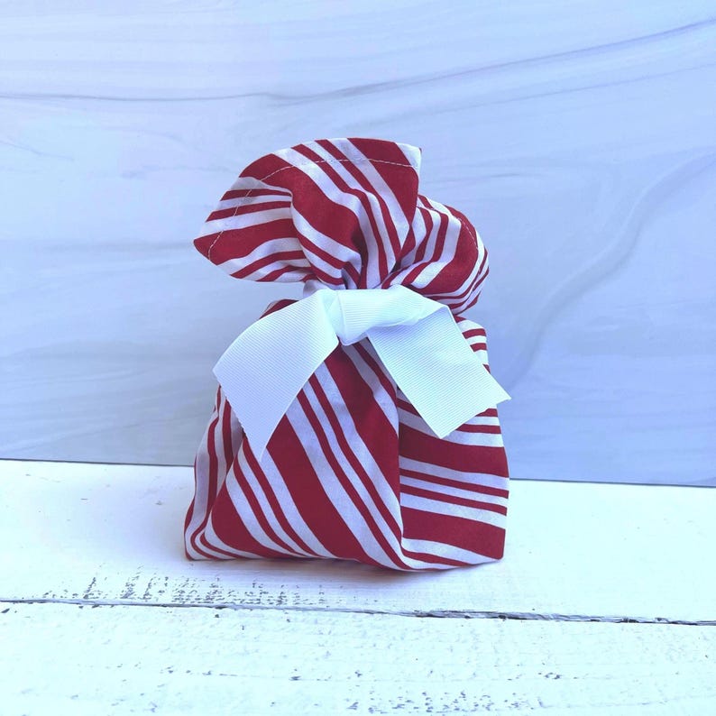 May include: A fabric drawstring pouch featuring a red and white diagonal striped pattern. The pouch is gathered at the top and secured with a crisp white grosgrain ribbon tied into a bow at the center, creating a festive and structured appearance.
