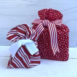 May include: Two fabric gift bags. The smaller bag features red and white diagonal stripes with a solid white ribbon bow. The larger bag is red with white polka dots, gathered at the top, and tied with a red and white striped fabric ribbon.