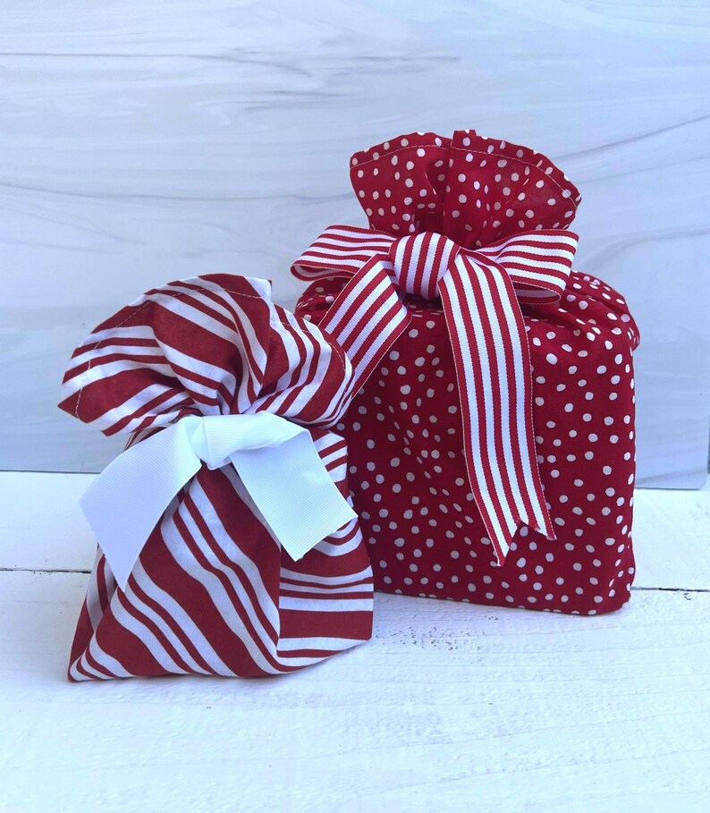 May include: Two fabric gift bags. The smaller bag features red and white diagonal stripes with a solid white ribbon bow. The larger bag is red with white polka dots, gathered at the top, and tied with a red and white striped fabric ribbon.