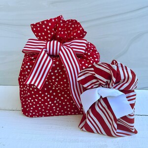 May include: Two fabric gift bags with drawstring closures. The larger bag is red with white polka dots and features a red and white striped ribbon bow. The smaller bag has a red and white striped pattern and a solid white ribbon bow.