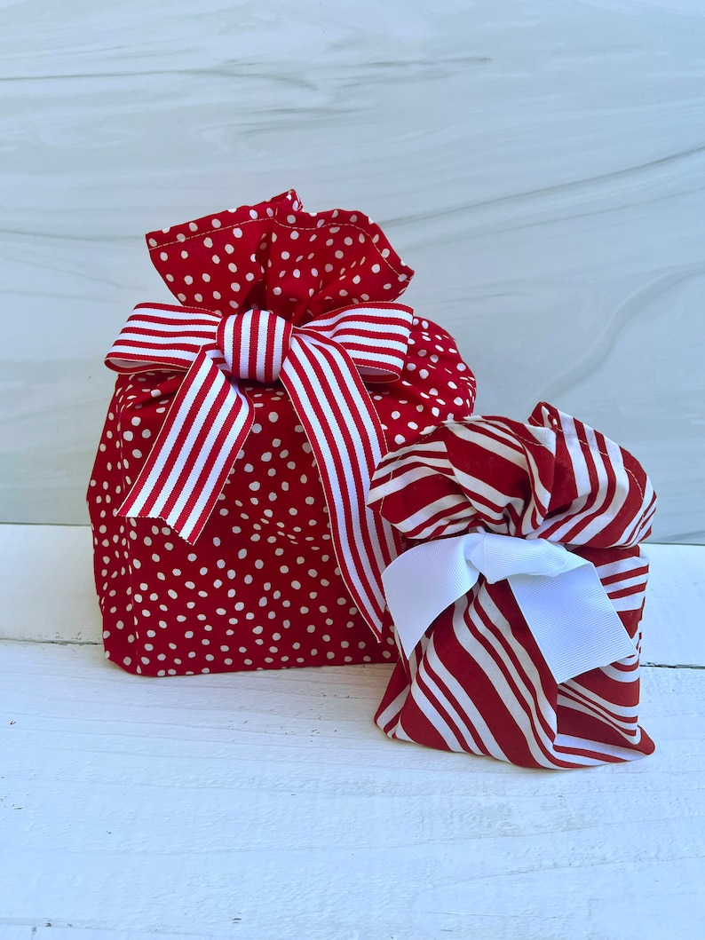 May include: Two fabric gift bags with drawstring closures. The larger bag is red with white polka dots and features a red and white striped ribbon bow. The smaller bag has a red and white striped pattern and a solid white ribbon bow.