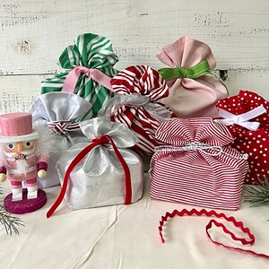 May include: A pink nutcracker figurine stands beside several fabric gift bags in various colors and patterns, including green and white stripes, red and white stripes, solid pink, silver, and red with white polka dots, all tied with ribbons.