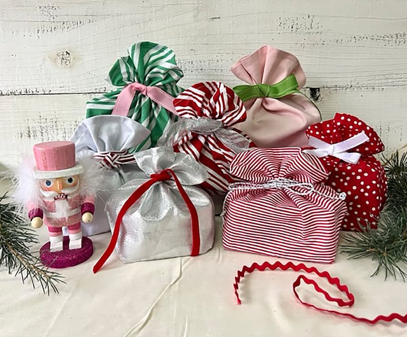 May include: A pink nutcracker figurine stands beside several fabric gift bags in various colors and patterns, including green and white stripes, red and white stripes, solid pink, silver, and red with white polka dots, all tied with ribbons.