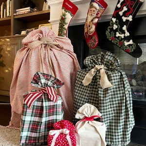 May include: A collection of five fabric gift bags in various sizes and patterns, including red and white stripes, green and white plaid, red and white polka dots, and solid cream. Each bag is cinched at the top with a tied fabric ribbon closure.