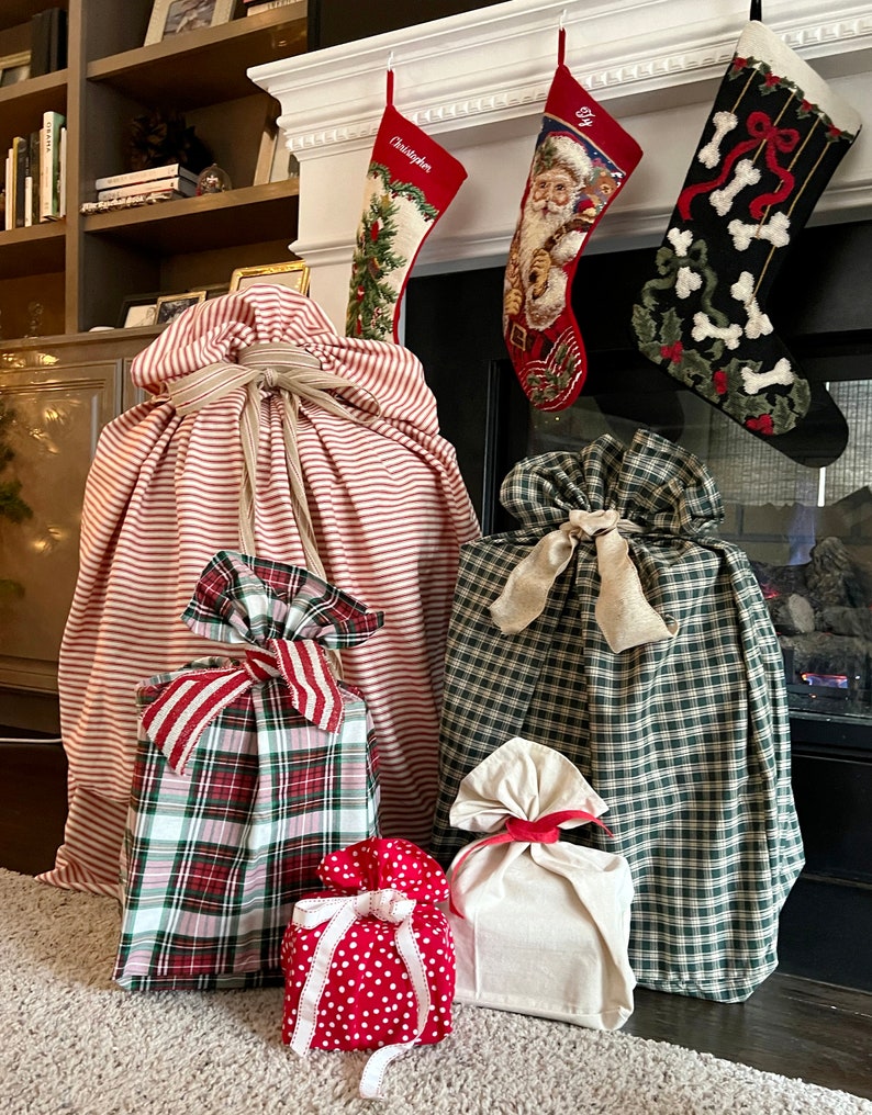 May include: A collection of five fabric gift bags in various sizes and patterns, including red and white stripes, green and white plaid, red and white polka dots, and solid cream. Each bag is cinched at the top with a tied fabric ribbon closure.