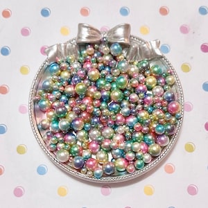 May include: A silver-colored, decorative dish filled with an assortment of iridescent pearls in various sizes. The pearls display a range of colors, including pink, green, blue, and yellow. A bow-shaped embellishment sits atop the dish.