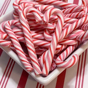 May include: A white square dish filled with red and white striped candy canes. The candy canes are curved and have a classic holiday design. The background features a red and white striped pattern.