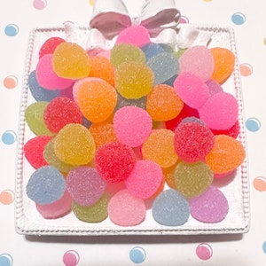 May include: A white rectangular tray filled with colorful, round candies. The candies are in shades of pink, orange, yellow, green, and blue. A white bow is at the top of the tray, and the background is a white surface with colorful polka dots.