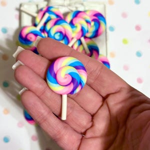 May include: A colorful, swirled lollipop in shades of pink, purple, blue, and yellow, held in a hand. The lollipop sits on a white stick. Other lollipops are visible in the background, along with a white box.