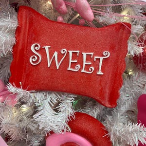 May include: A red, candy-shaped ornament with the word "SWEET" in white script. The ornament is part of a festive display, surrounded by white and pink decorations, including a large, red swirl and pink, oblong shapes.