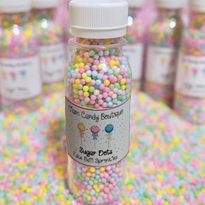 May include: A clear plastic bottle filled with colorful sugar dots, with a white cap. The label reads "Glam Candy Boutique" and "Sugar Dots Cake Ball Sprinkles". The background is filled with similar sprinkles.