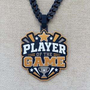 Puede incluir: Un collar con colgante azul marino y dorado que presenta un diseño en forma de escudo con el texto "PLAYER OF THE GAME" en letras blancas y doradas. El colgante incluye una estrella central, un trofeo y una cadena gruesa.