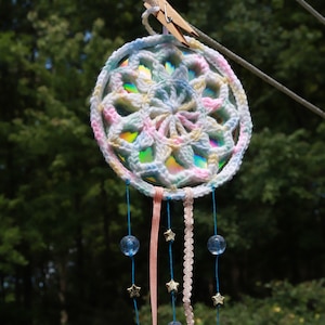 Puede incluir: Un atrapasueños de crochet blanco con detalles en rosa pastel, amarillo y azul cuelga de un tendedero. Tiene cuentas de vidrio azules y abalorios de estrellas doradas en cuerdas azules.