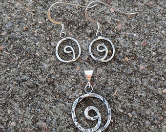 Puede incluir: Un conjunto de pendientes y un collar con colgante de plata con un diseño en espiral. Los pendientes son simples y delicados, mientras que el colgante es más elaborado con un acabado texturizado.