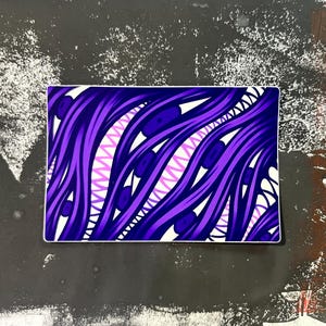 May include: A rectangular sticker with a vibrant abstract design. The artwork features flowing, wavy lines in shades of purple and white, with a central zig-zag pattern in light pink. Dark blue accents and white shapes add depth to the design.
