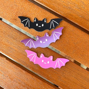 May include: Three bat-shaped stickers in black, purple, and pink, each with a smiling face and white wing details. The stickers are arranged on a wooden surface with metal accents, creating a playful Halloween theme.