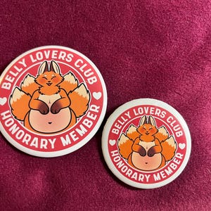 Peut inclure: Deux badges ronds blancs avec une bordure rouge. Ils présentent un renard de dessin animé et le texte "Belly Lovers Club Honorary Member". Le renard a une fourrure orange et un ventre rose clair.