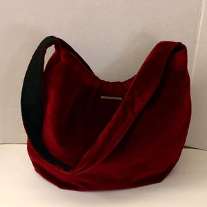 May include: A burgundy velvet shoulder bag with a black strap. The bag has a curved shape and a small rectangular tag near the top. The bag is made of a soft, plush material.