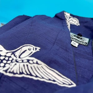 May include: A navy blue garment with a white bird design. The garment has a tag that reads "sannah" and "ONE SIZE". The bird design is on the front and the collar of the garment. The background is a bright blue.