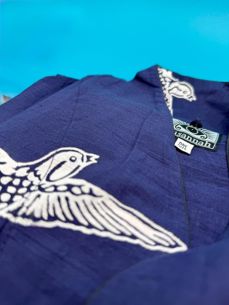 May include: A navy blue garment with a white bird design. The garment has a tag that reads "sannah" and "ONE SIZE". The bird design is on the front and the collar of the garment. The background is a bright blue.
