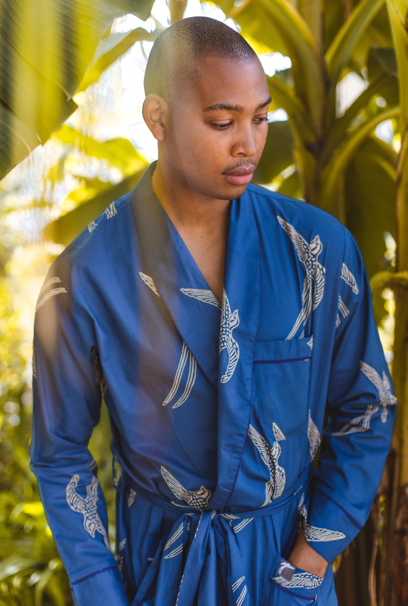 May include: A blue robe with a bird pattern. The robe has a shawl collar, a tie belt, and a pocket. The bird pattern is white and beige. The robe is made of a soft, flowing fabric.