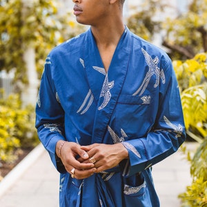 May include: A blue robe with a white bird pattern. The robe has a shawl collar, a front pocket, and a matching belt. The sleeves are long, and the robe is made of a soft, flowing fabric. The model is wearing gold rings and a bracelet.