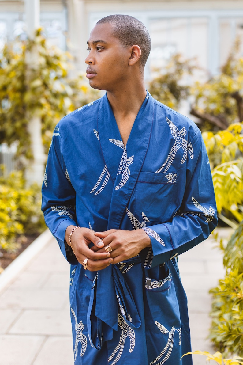 May include: A blue robe with a white bird pattern. The robe has a shawl collar, a front pocket, and a matching belt. The sleeves are long, and the robe is made of a soft, flowing fabric. The model is wearing gold rings and a bracelet.