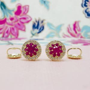 May include: A pair of gold-colored stud earrings with a central cluster of deep red gemstones surrounded by a halo of smaller, clear stones. The earrings are set against a soft pink background.