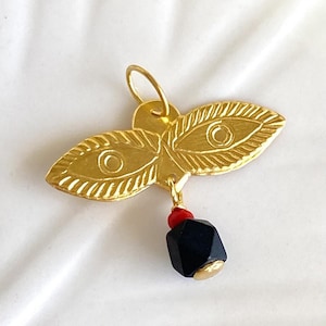 May include: A gold-colored pendant with a leaf-like design, featuring eye-like details. A red bead and a black, geometric bead hang below. The pendant is on a white surface.