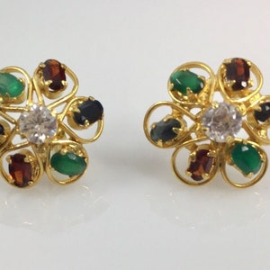 May include: A pair of gold earrings with a flower design. Each earring has a white center stone surrounded by six smaller stones in various colors, including green, blue, and red.